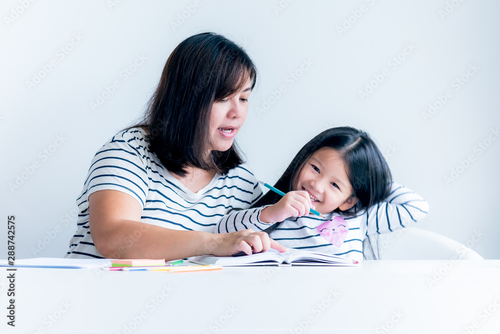 An Asian mother is teaching a 6 year old daughter to learn to write and read On white table and white wall background, concept to family and education. An Asian mother is teaching a 6 year old daughter to learn to write and read On white table and white wall background, concept to family and education.