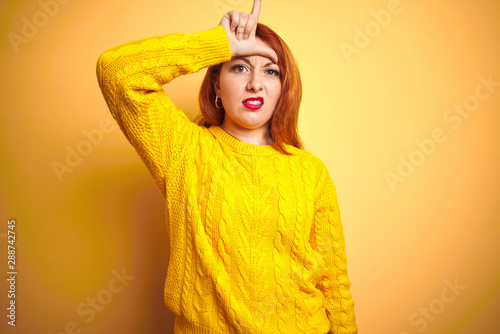 Beautiful redhead woman wearing winter sweater standing over isolated yellow background making fun of people with fingers on forehead doing loser gesture mocking and insulting. © Krakenimages.com