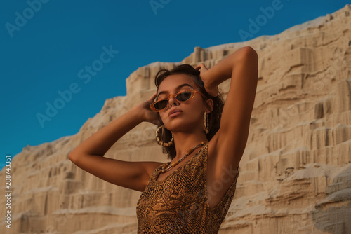 Outdoor portrait of young beautiful woman with luxury tanned skin wearing snakeskin print swimwear, trendy sunglasses, hoop earrings, posing in sand desert, at sunset. Copy, empty space for text  photo