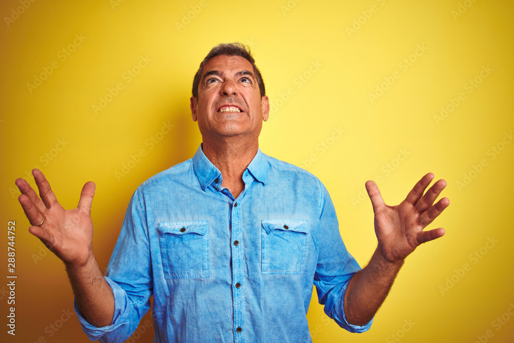 Handsome middle age man wearing denim shirt standing over isolated yellow background crazy and mad shouting and yelling with aggressive expression and arms raised. Frustration concept. Handsome middle age man wearing denim shirt standing over isolated yellow background crazy and mad shouting and yelling with aggressive expression and arms raised. Frustration concept.