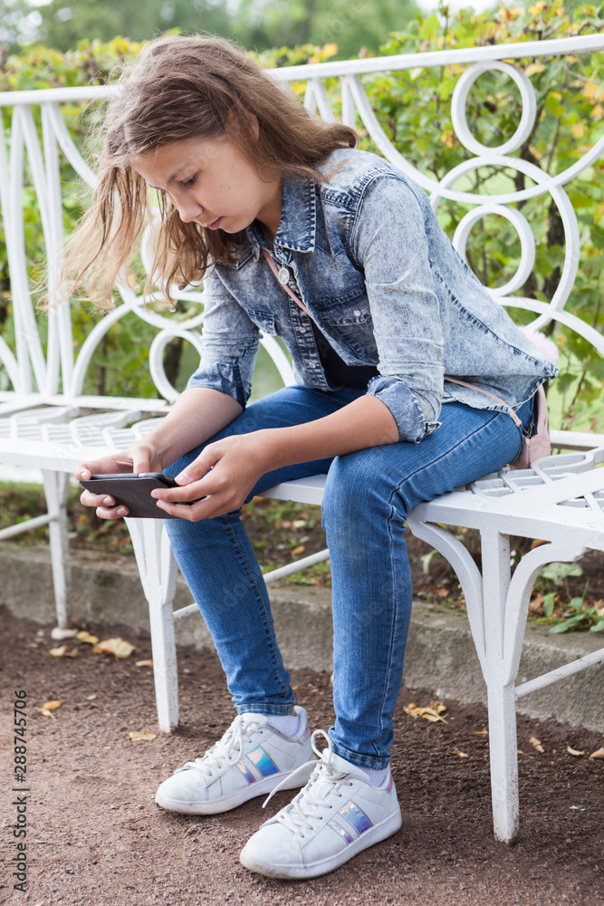 Attractive Caucasian teen age girl using smartphone while sitting on bench  in park, jeans modern clothing Stock Photo | Adobe Stock