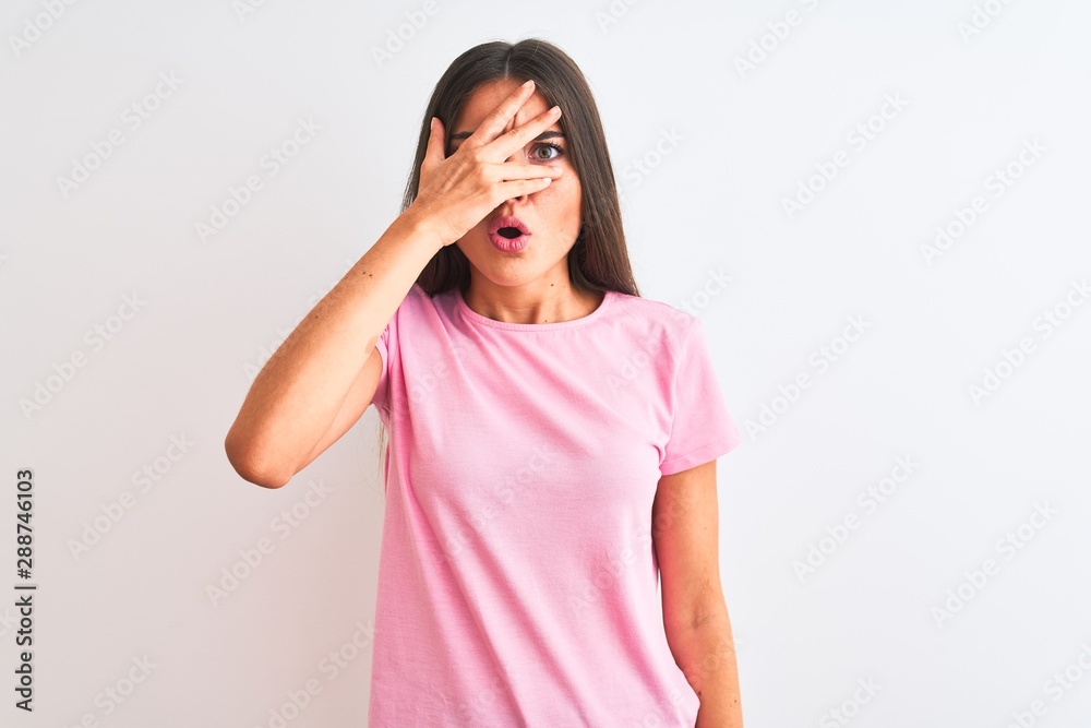 Young beautiful woman wearing pink casual t-shirt standing over isolated white background peeking in shock covering face and eyes with hand, looking through fingers with embarrassed expression. Young beautiful woman wearing pink casual t-shirt standing over isolated white background peeking in shock covering face and eyes with hand, looking through fingers with embarrassed expression.