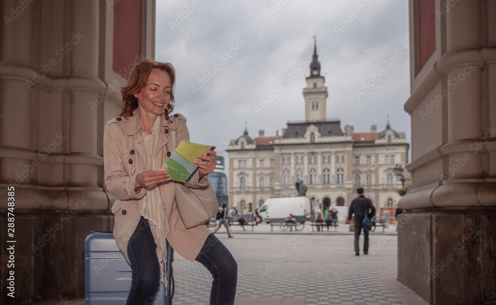 One smiling mature woman, walking in a pedestrian zone, in a city. Traveler pushing her suitcase.