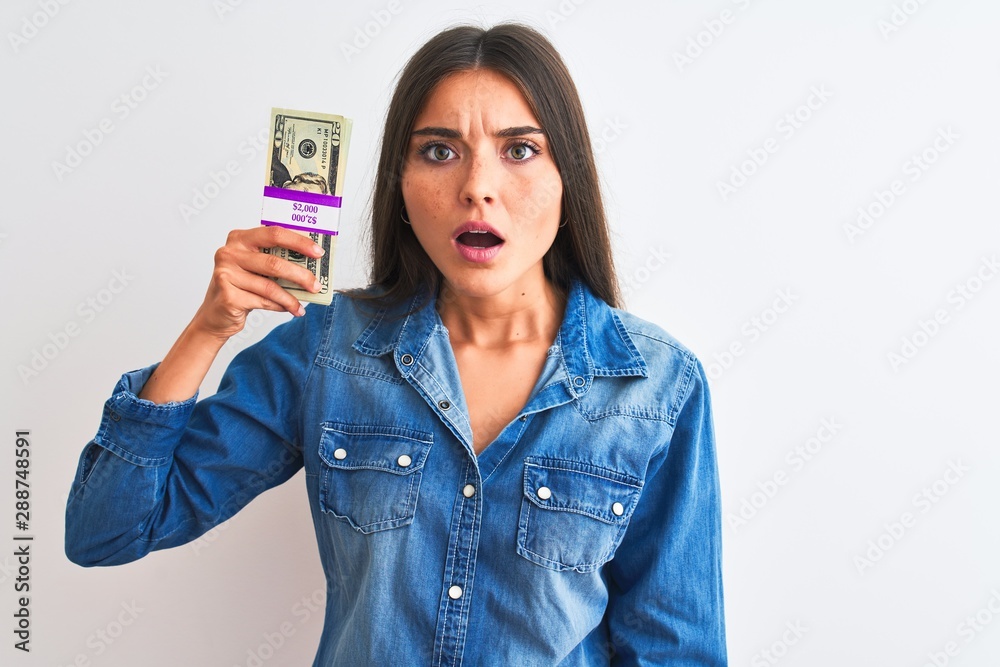 Young beautiful woman holding dollars standing over isolated white background scared in shock with a surprise face, afraid and excited with fear expression Young beautiful woman holding dollars standing over isolated white background scared in shock with a surprise face, afraid and excited with fear expression