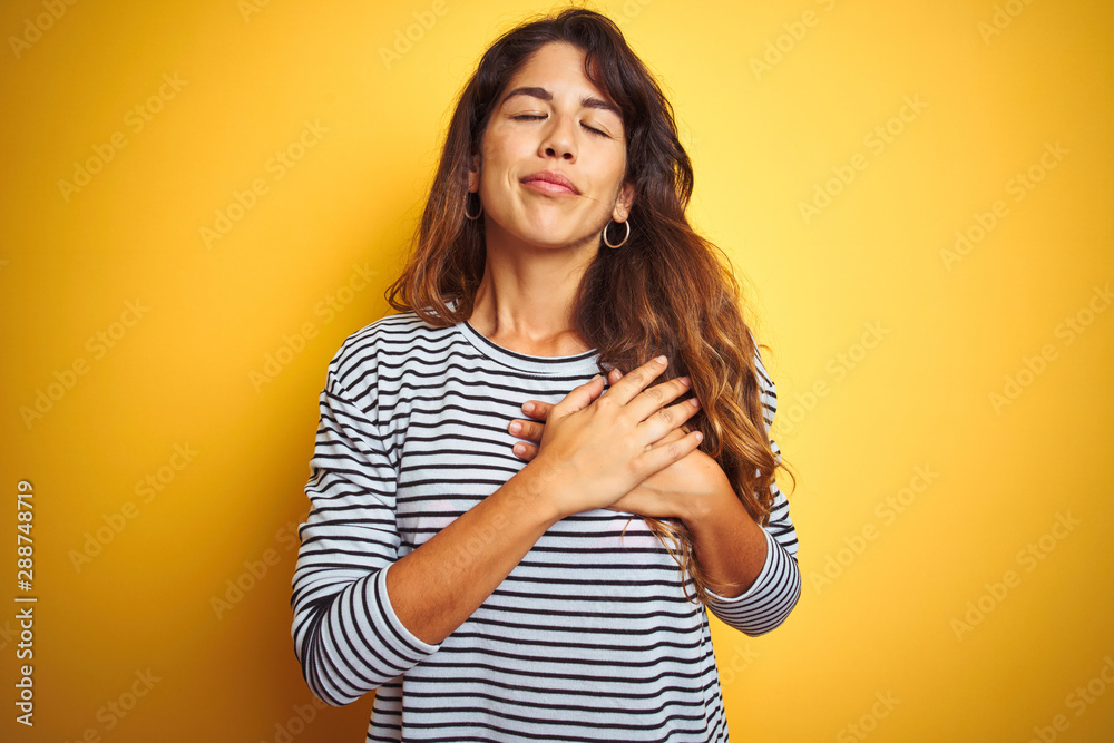 Young beautiful woman wearing stripes t-shirt standing over yelllow isolated background smiling with hands on chest with closed eyes and grateful gesture on face. Health concept.