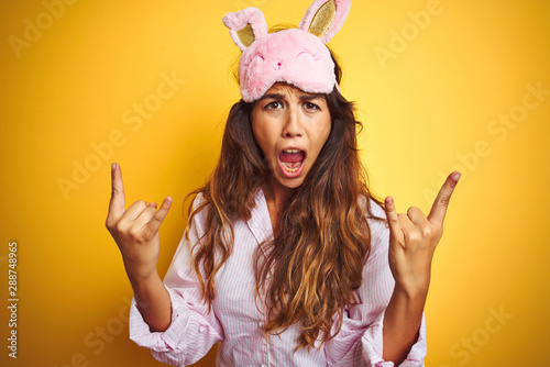 Young woman wearing pajama and sleep mask standing over yellow isolated background shouting with crazy expression doing rock symbol with hands up. Music star. Heavy music concept. photo