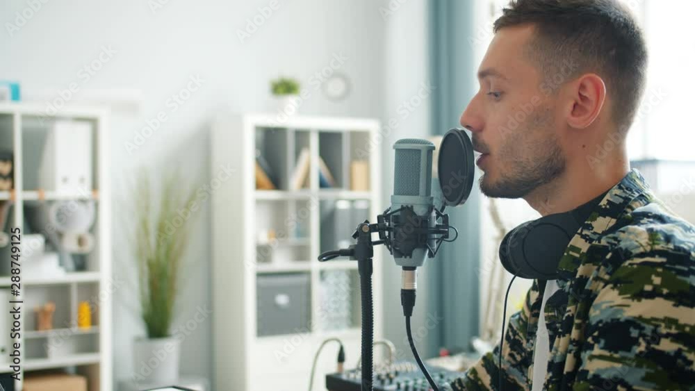 Attractive young guy successful blogger is speaking in microphone recording podcast for online blog working in apartment. People and mass media concept. Attractive young guy successful blogger is speaking in microphone recording podcast for online blog working in apartment. People and mass media concept.