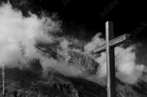 Close up plain wooden summit cross (Jesus Christ crucifix) with the Alps mountain ranges, low hang thick clouds and dramatic dark black sky. Black and white image for Halloween or spiritual concept.