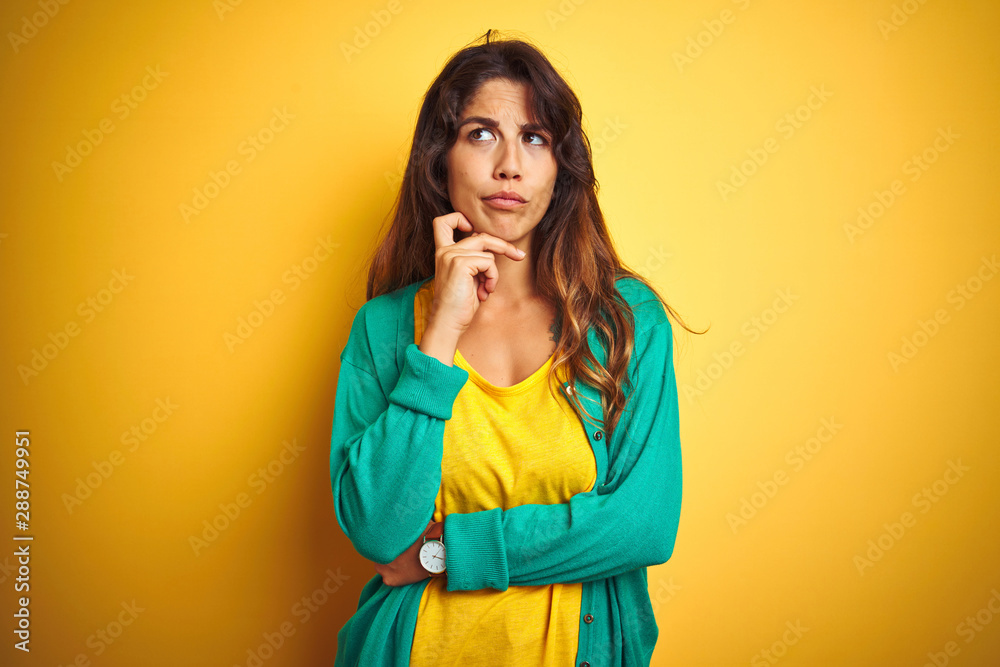 Young woman wearing t-shirt and green sweater standing over yelllow isolated background with hand on chin thinking about question, pensive expression. Smiling with thoughtful face. Doubt concept. Young woman wearing t-shirt and green sweater standing over yelllow isolated background with hand on chin thinking about question, pensive expression. Smiling with thoughtful face. Doubt concept.