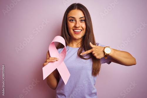 Young beautiful woman holding cancer ribbon standing over isolated pink background with surprise face pointing finger to himself