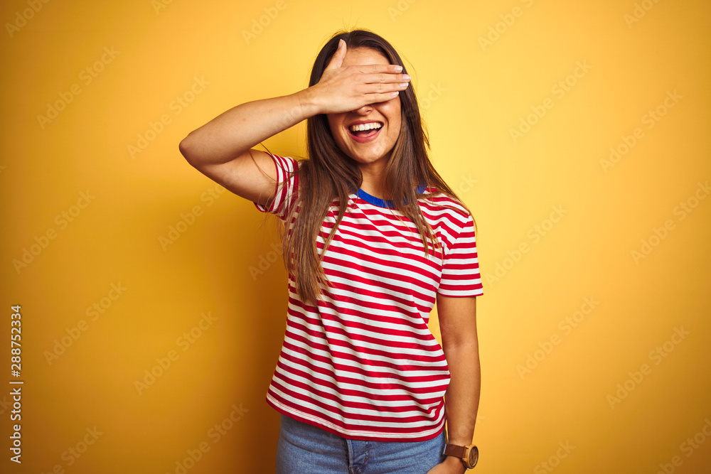 Young beautiful woman wearing striped t-shirt standing over isolated yellow background smiling and laughing with hand on face covering eyes for surprise. Blind concept. Young beautiful woman wearing striped t-shirt standing over isolated yellow background smiling and laughing with hand on face covering eyes for surprise. Blind concept.