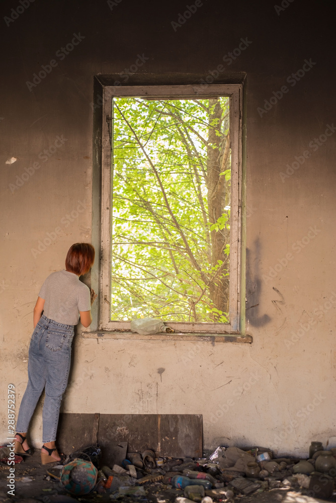 Fototapeta premium girl in a dirty room looks out of the window at green trees. Environmental pollution. Changing of the climate.