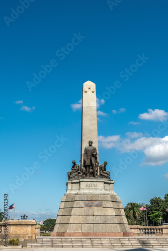 Manila, Philippines - March 5, 2019: Obelisk with bronze statues of Jose Rizal stands on brown stone pedestal under blue sky in Rizal Park. Flags and green foliage in back. photo