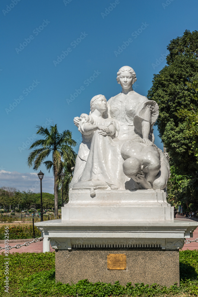 Manila, Philippines - March 5, 2019: Closeup of white La Madre Filipina statue in Rizal Park under blue sky with green foliage.