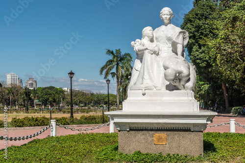 Manila, Philippines - March 5, 2019: Wide shot of white La Madre Filipina statue in Rizal Park under blue sky with green foliage. Some cityscape.