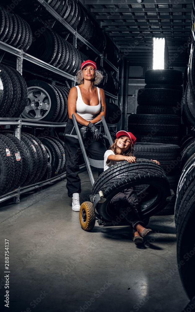 Strong female worker and her little helper have fun while looking for right tyres at warehouse. - obrazy, fototapety, plakaty 