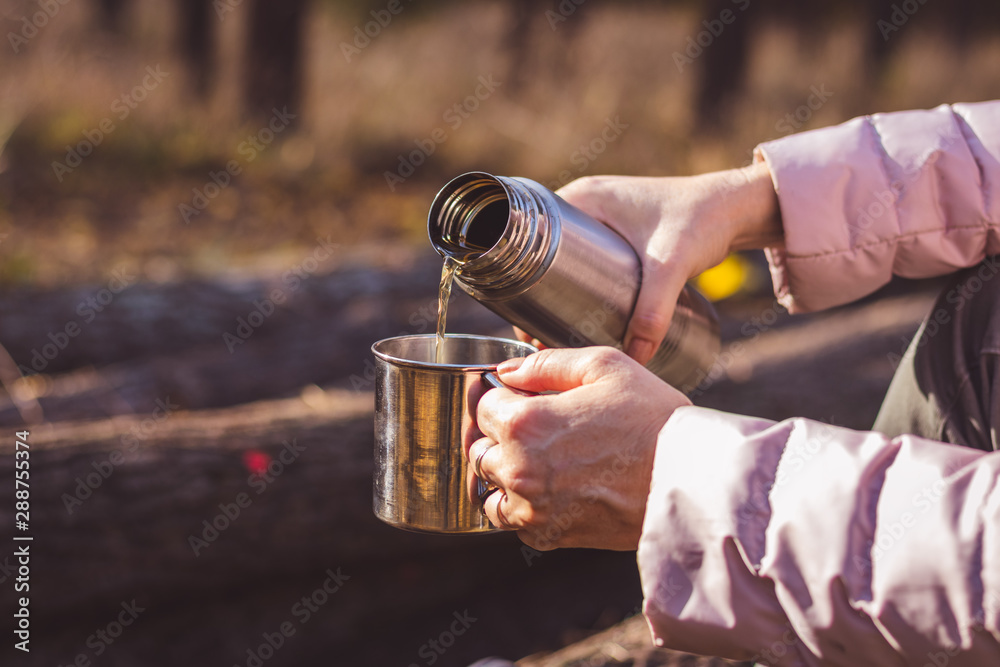 Woman pouring hot drink out of thermos into metal cup in autumn forest Woman pouring hot drink out of thermos into metal cup in autumn forest
