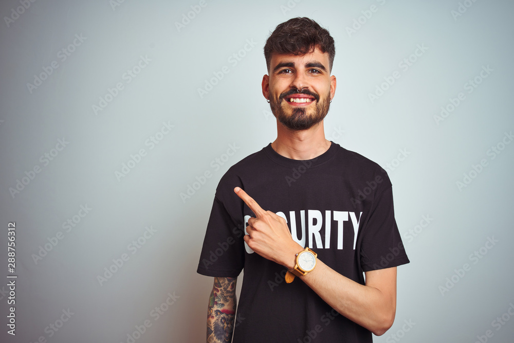 Young safeguard man with tattoo wering security uniform over isolated white background cheerful with a smile of face pointing with hand and finger up to the side with happy and natural