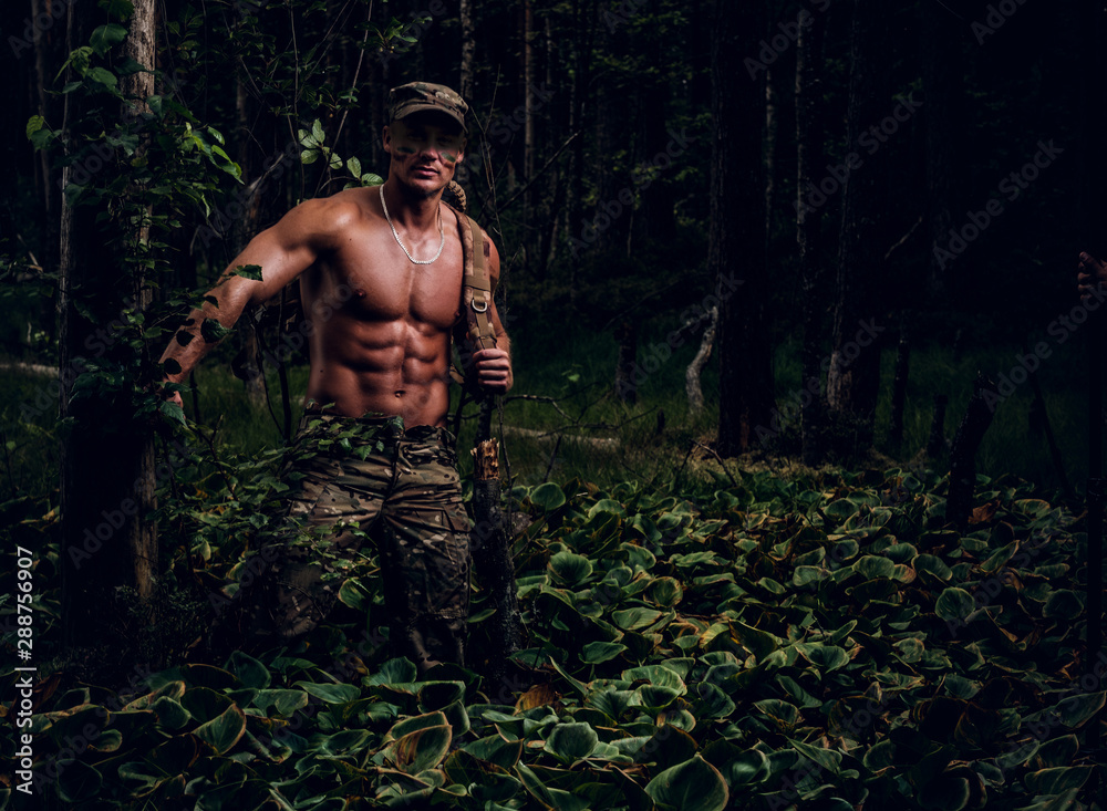Brutal muscular man in military uniform and naked torso is posing for a photographer in the forest.