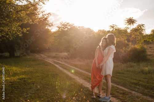Two girlfriends walk together in the woods at sunset. It is beautiful summer weather. Both are dressed in dresses.