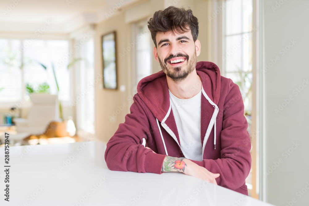 Young man wearing casual sweatshirt sitting on white table happy face smiling with crossed arms looking at the camera. Positive person.