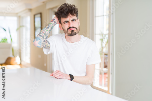 Young man wearing casual t-shirt sitting on white table confuse and wonder about question. Uncertain with doubt, thinking with hand on head. Pensive concept.