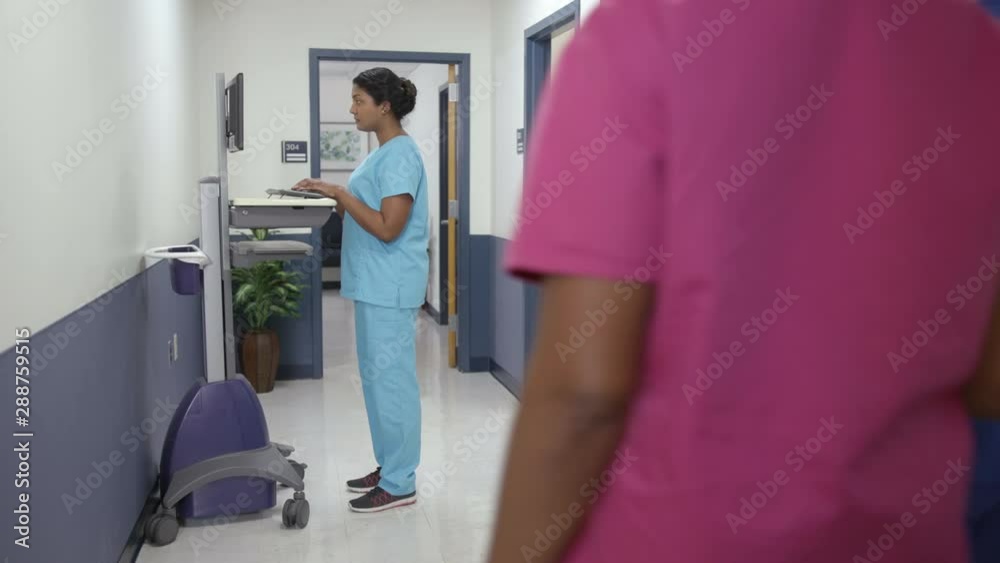 Hospital hallway, nurse types on EMR cart as nurses pass camera moves left right