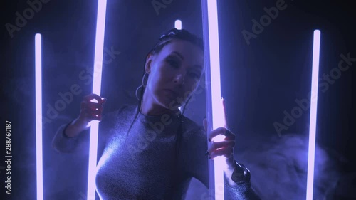 Neon night. Fashionable girl in stylish shiny dress among neon light tubes in a warm room. Girl of Asian appearance. Big earrings, hairstyle braids. Punk style. Smoke and blue neon.