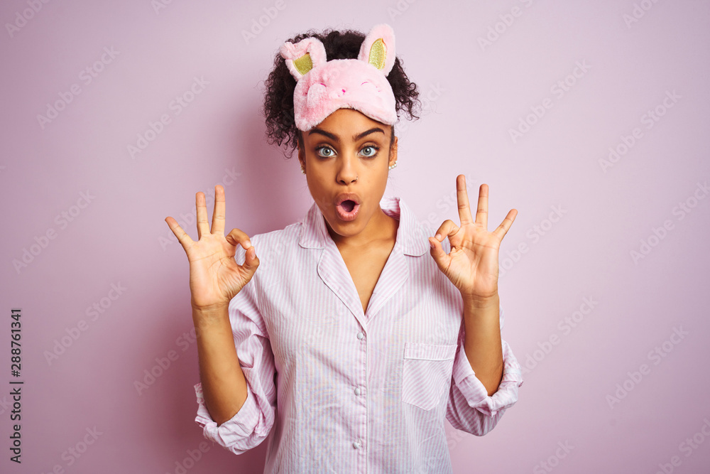 Young african american woman wearing pajama and mask over isolated pink background looking surprised and shocked doing ok approval symbol with fingers. Crazy expression - obrazy, fototapety, plakaty 