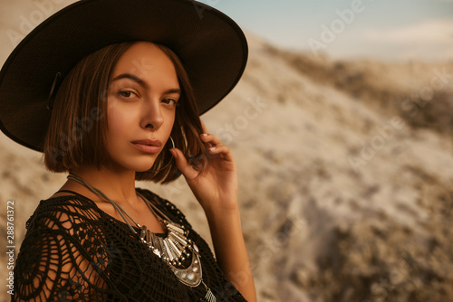 Outdoor close up portrait of young beautiful woman with tanned skin wearing black hat, boho, ethnic necklace, crochet top, posing on sand, at sunset. Copy, empty space for text 