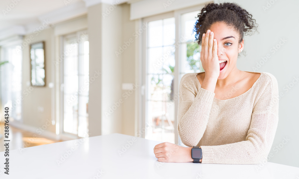 Beautiful young african american woman with afro hair sitting on table at home covering one eye with hand with confident smile on face and surprise emotion. Beautiful young african american woman with afro hair sitting on table at home covering one eye with hand with confident smile on face and surprise emotion.