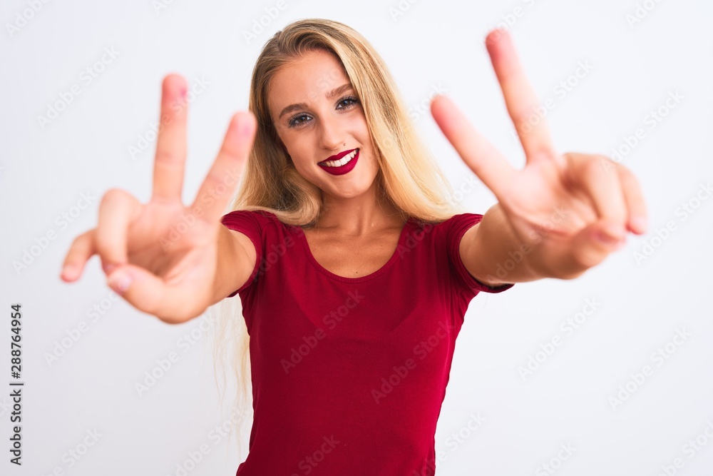 Young beautiful woman wearing red t-shirt standing over isolated white background smiling looking to the camera showing fingers doing victory sign. Number two. Young beautiful woman wearing red t-shirt standing over isolated white background smiling looking to the camera showing fingers doing victory sign. Number two.