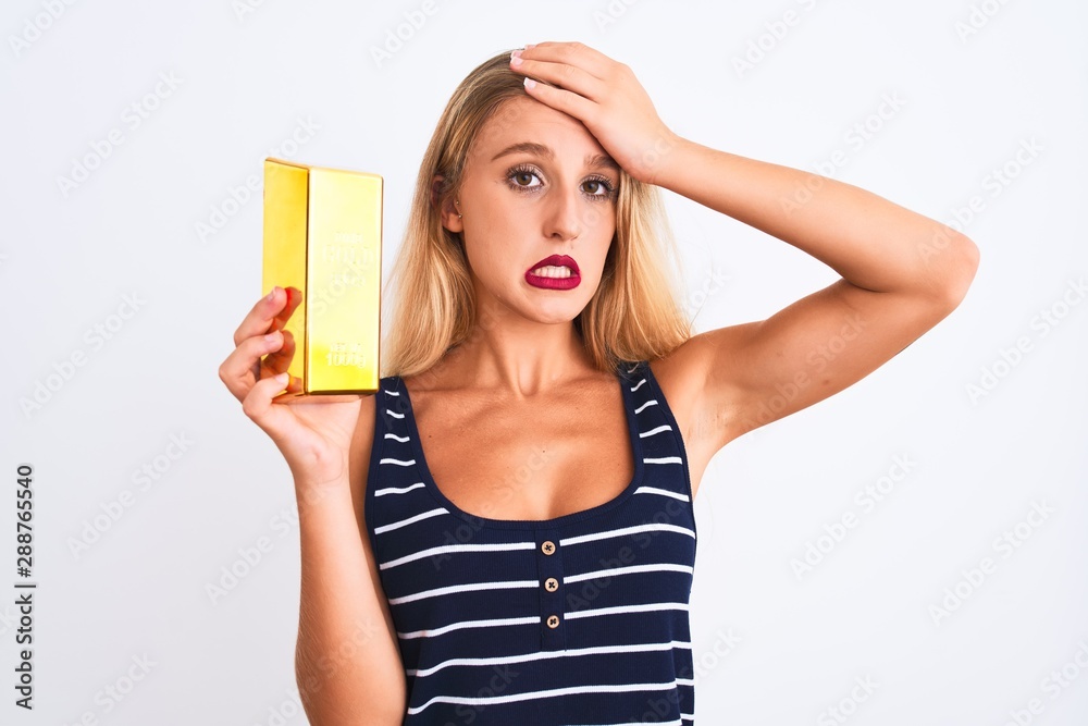 Young beautiful blond woman holding gold ingot standing over isolated white background stressed with hand on head, shocked with shame and surprise face, angry and frustrated Young beautiful blond woman holding gold ingot standing over isolated white background stressed with hand on head, shocked with shame and surprise face, angry and frustrated