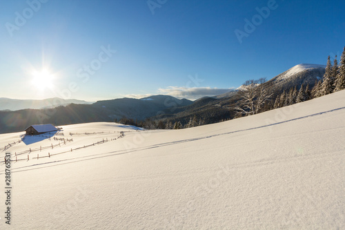Winter Christmas landscape of mountain valley on frosty sunny day. Old wooden forsaken shepherd hut in white deep clean snow, woody dark mountain ridge, bright sun on blue sky copy space background.