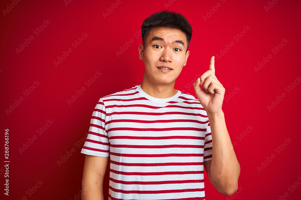 Fototapeta premium Young asian chinese man wearing striped t-shirt standing over isolated red background pointing finger up with successful idea. Exited and happy. Number one.