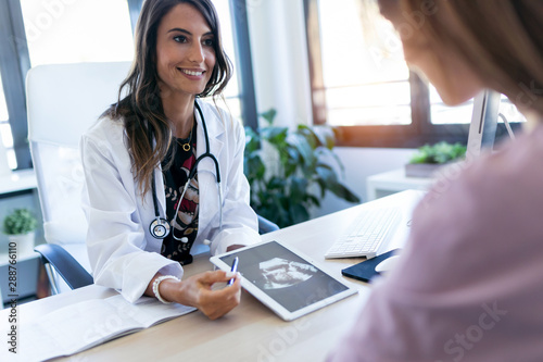 Pretty young woman gynecologist using digital tablet for showing the ultrasound to her pregnant patient in the clinic. photo