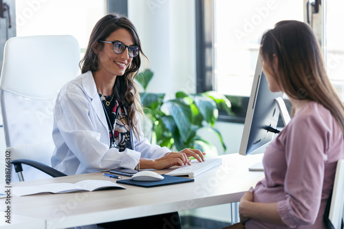 Foto Pretty young woman gynecologist reviewing the medical history of her pregnant patient with computer in the clinic