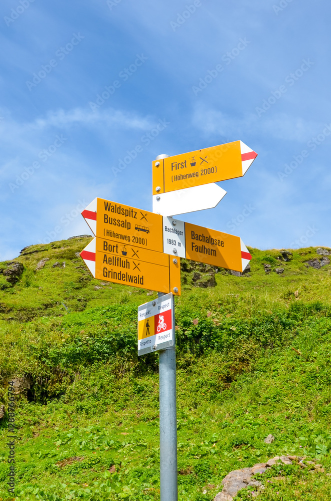 Yellow tourist sign in Bachlager, Switzerland giving distances and directions to hikers in the Swiss Alps. Popular hiking trail by Grindelwald leading to Bachalpsee. Alpine stream in the background