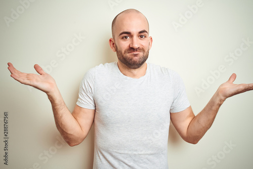 Young bald man with beard wearing casual white t-shirt over isolated background clueless and confused expression with arms and hands raised. Doubt concept.