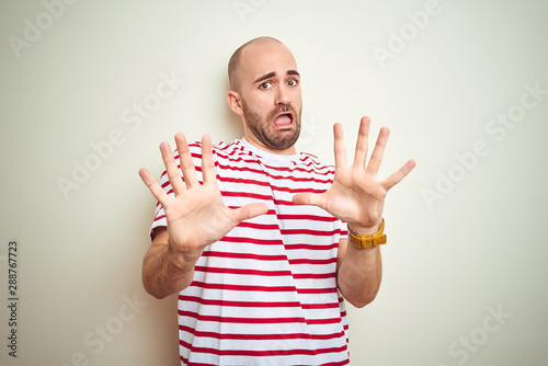 Young bald man with beard wearing casual striped red t-shirt over white isolated background afraid and terrified with fear expression stop gesture with hands, shouting in shock. Panic concept.