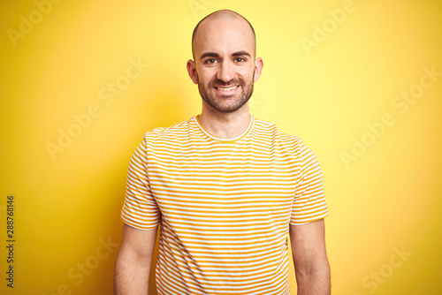 Young bald man with beard wearing casual striped t-shirt over yellow isolated background with a happy and cool smile on face. Lucky person.