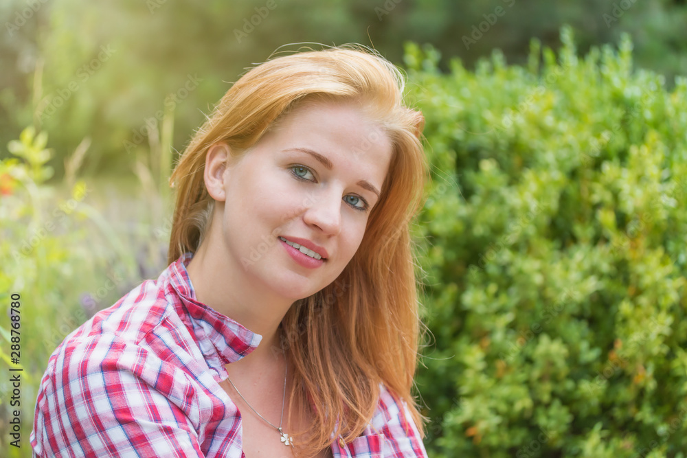 Attractive young woman clothed in checkered shirt is smiling, showing her white teeth looking at the camera outdoors.