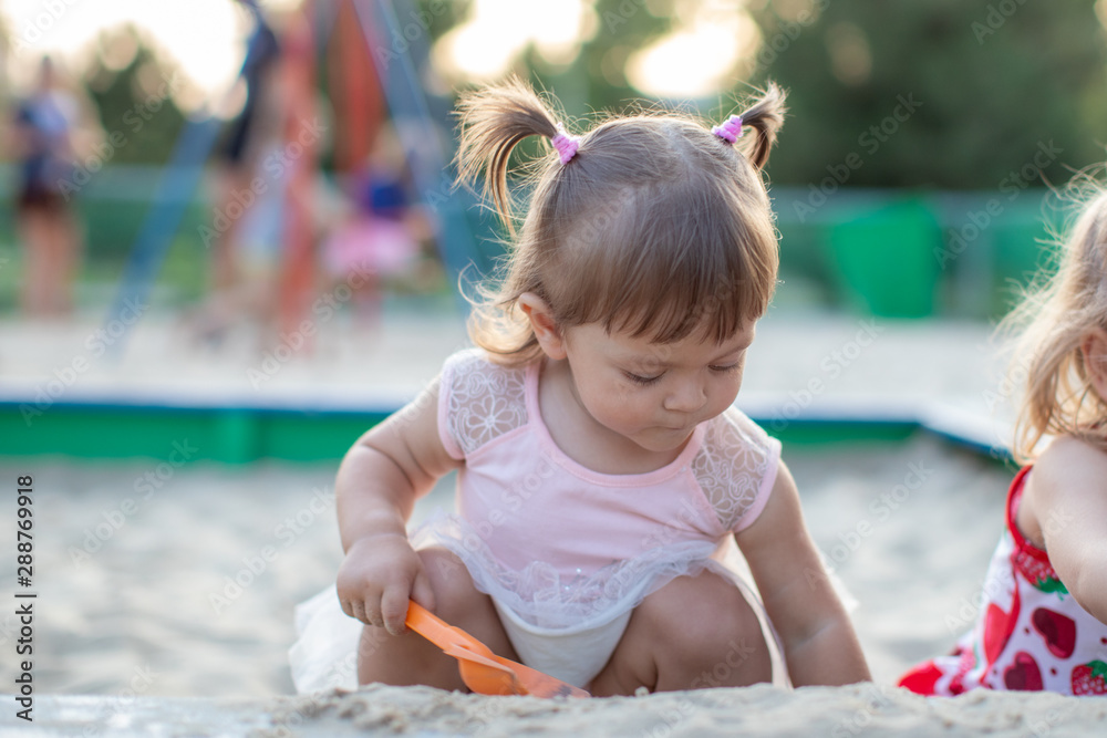 Naklejka premium Cute toddler girl playing in sand on outdoor playground. Beautiful baby in red trousers having fun on sunny warm summer day. Child with colorful sand toys. Healthy active baby outdoors plays games