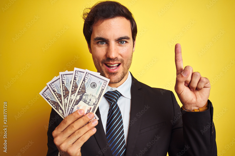 Young handsome businessman wearing suit holding dollars over isolated yellow background surprised with an idea or question pointing finger with happy face, number one Young handsome businessman wearing suit holding dollars over isolated yellow background surprised with an idea or question pointing finger with happy face, number one