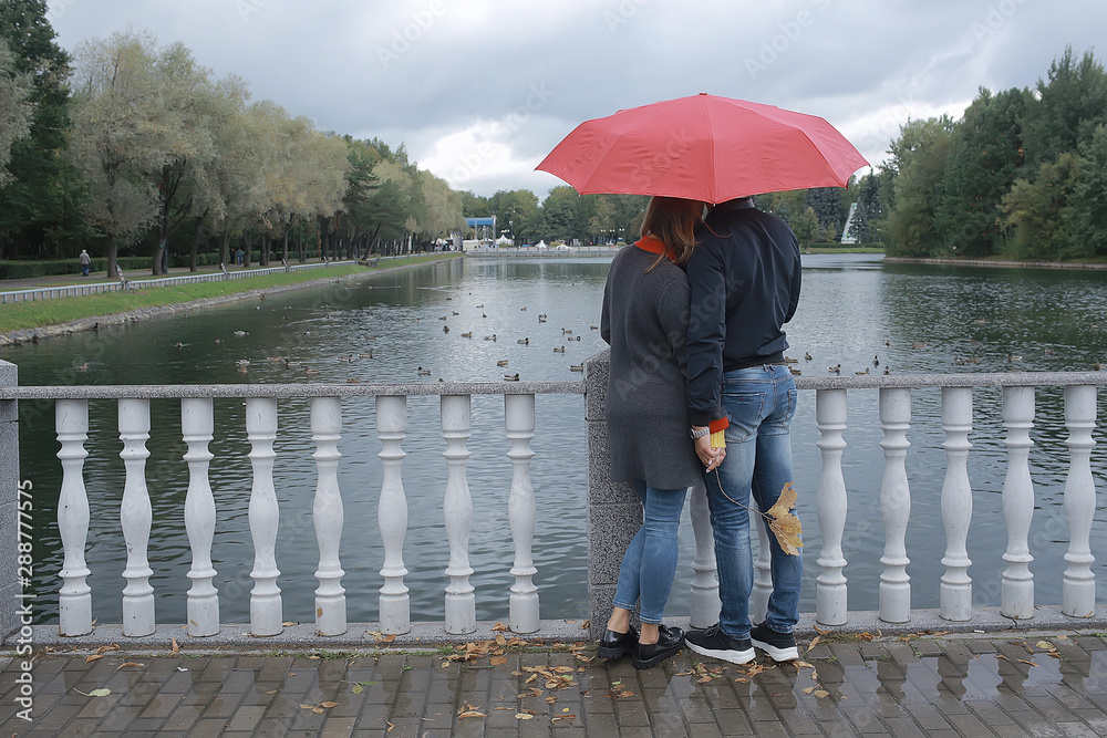 Obraz premium rain in the autumn park / young 25 years old couple man and woman walk under an umbrella in wet rainy weather, walk October lovers