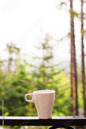 White coffee cup on the balcony with blurred nature view and forest background, after rain