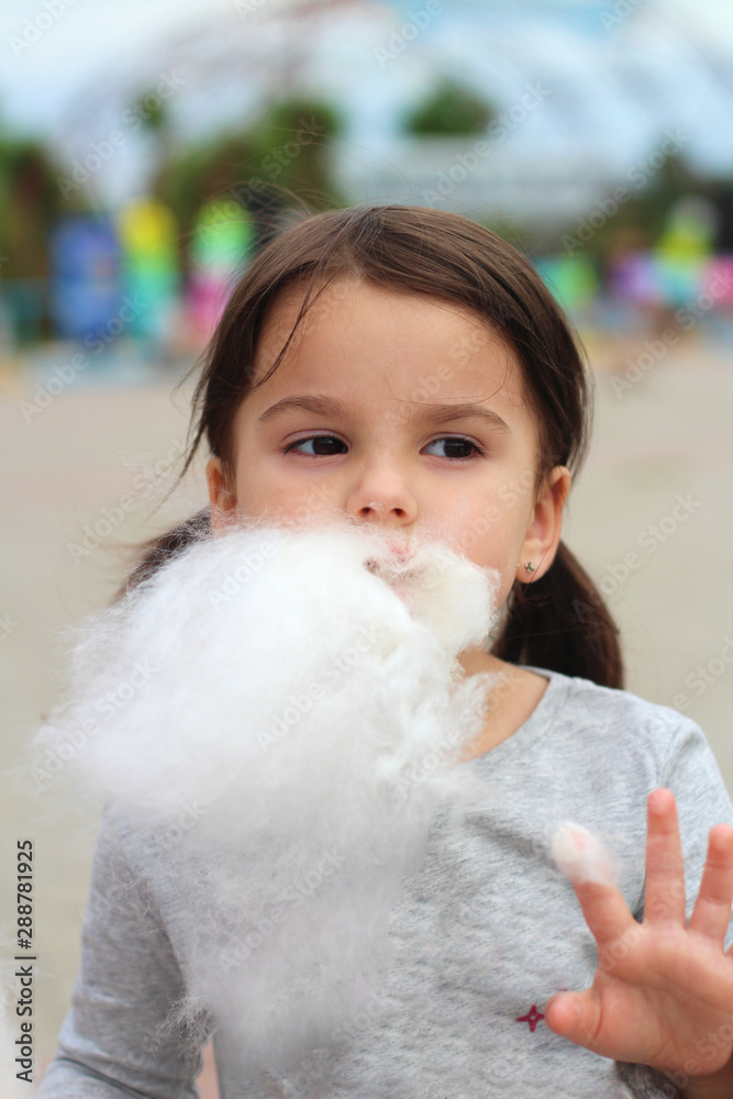 Little funny white girl with tails with narrow eyes holds cotton candy with his lips while walking outdoors Little funny white girl with tails with narrow eyes holds cotton candy with his lips while walking outdoors