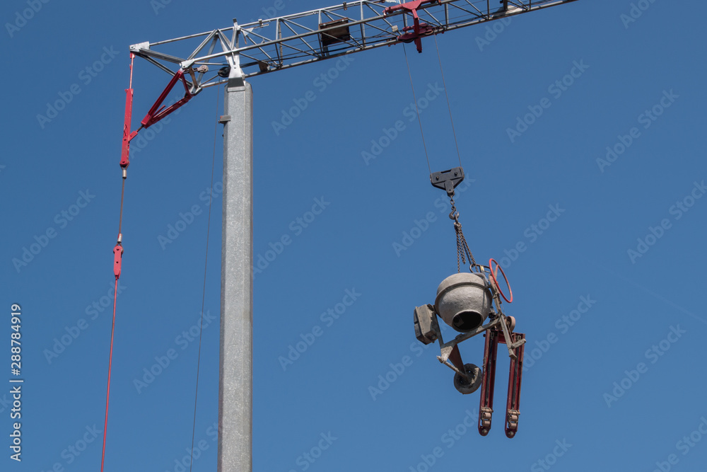 A round grey concrete mixer and a red movable carriage trolley are suspended from the boom hook of a tower crane with a grey vertical column and a red counterweight A round grey concrete mixer and a red movable carriage trolley are suspended from the boom hook of a tower crane with a grey vertical column and a red counterweight