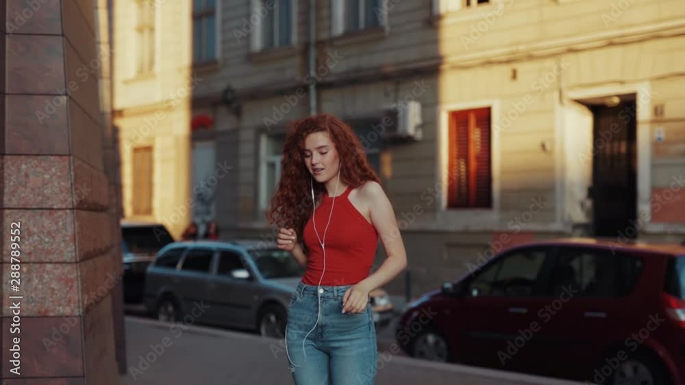 Pretty curly young girl is having fun while walking around the city and dancing. Student girl listening to music via white headphones and happily walking around. Facial expression, positive emotions Pretty curly young girl is having fun while walking around the city and dancing. Student girl listening to music via white headphones and happily walking around. Facial expression, positive emotions