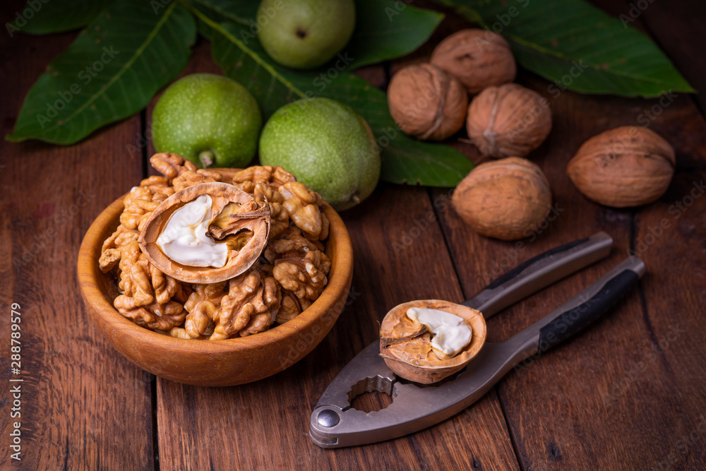 Walnuts chopped in a bowl, whole and in a green peel and a nut cracker on a dark old wooden background.